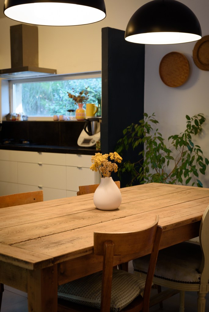 Une cuisine moderne avec une grande table en bois, des chaises, un vase de fleurs séchées, et des luminaires noirs suspendus.