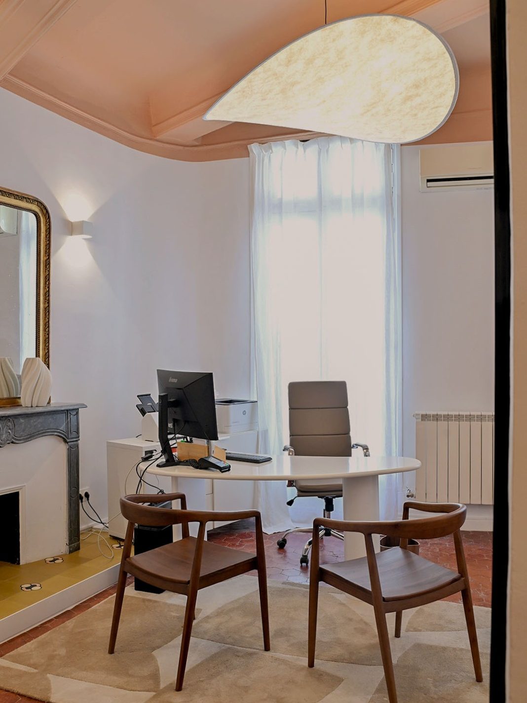 Bureau moderne avec une lumière suspendue en forme de goutte, un ordinateur sur un bureau rond, deux chaises en bois, et une fenêtre avec des rideaux blancs.