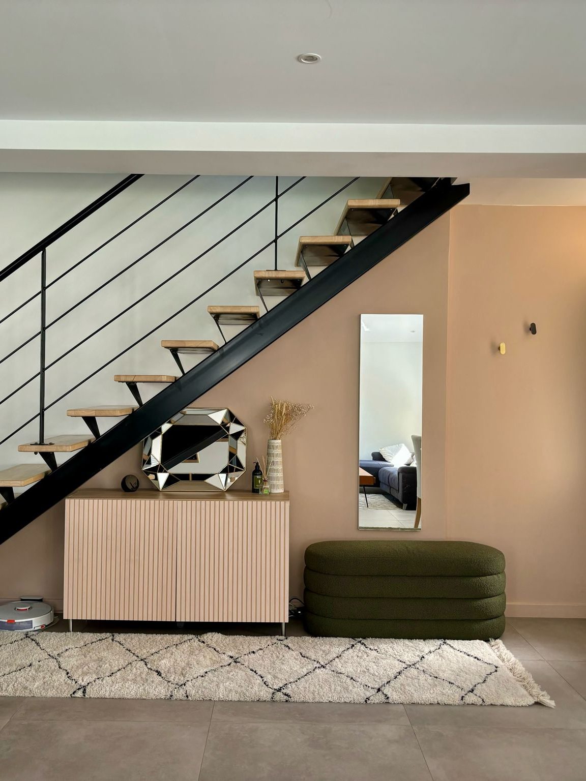 Vue d'un hall d'entrée moderne avec un escalier en bois et métal, un meuble en bois clair avec un miroir décoratif, un vase de fleurs sèches et un coussin vert sur le sol.