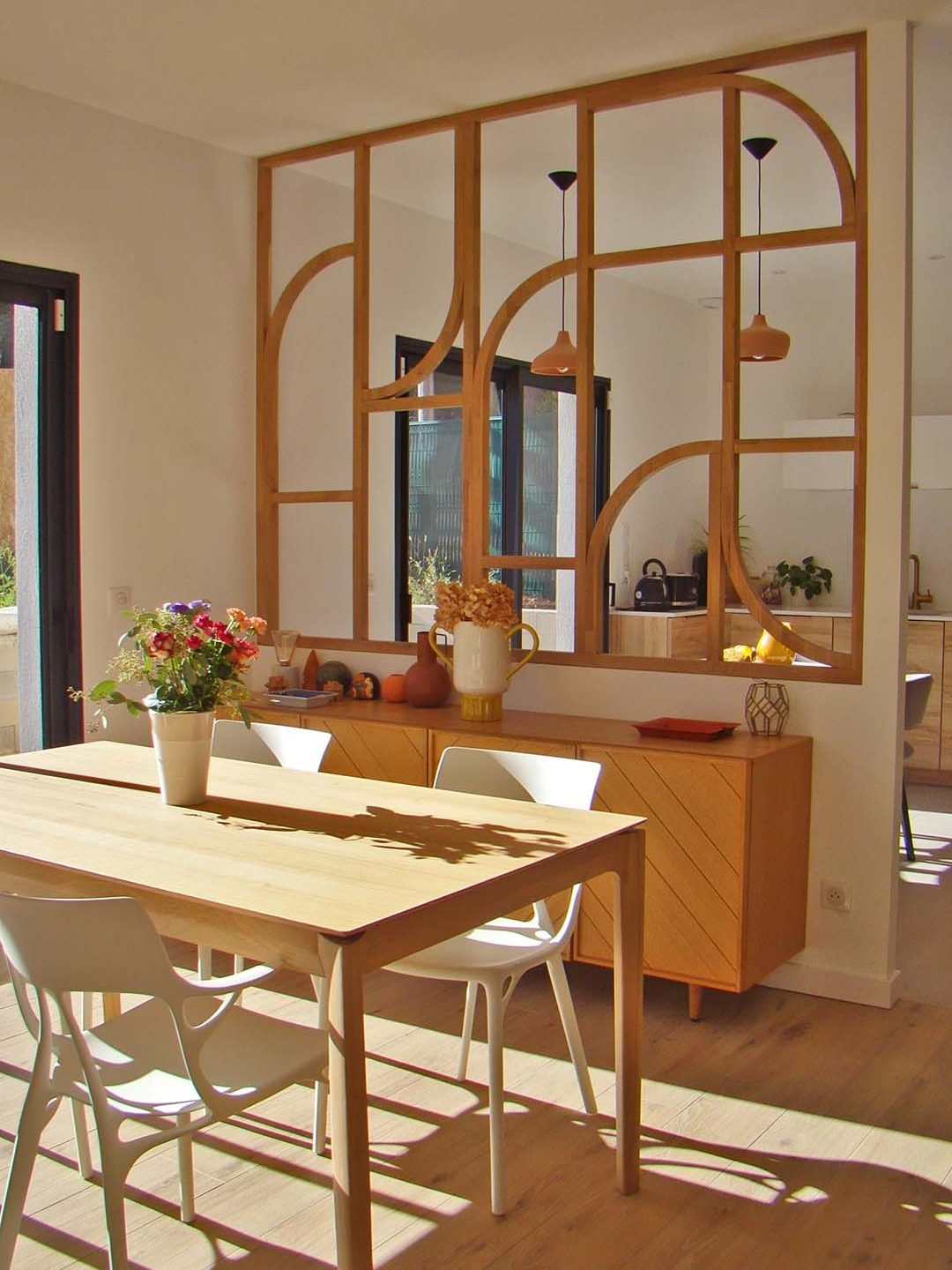 Une cuisine moderne avec une table en bois, des chaises blanches et un rangement en bois. Un mur de séparation en bois design avec des fenêtres et des suspensions lumineuses.
