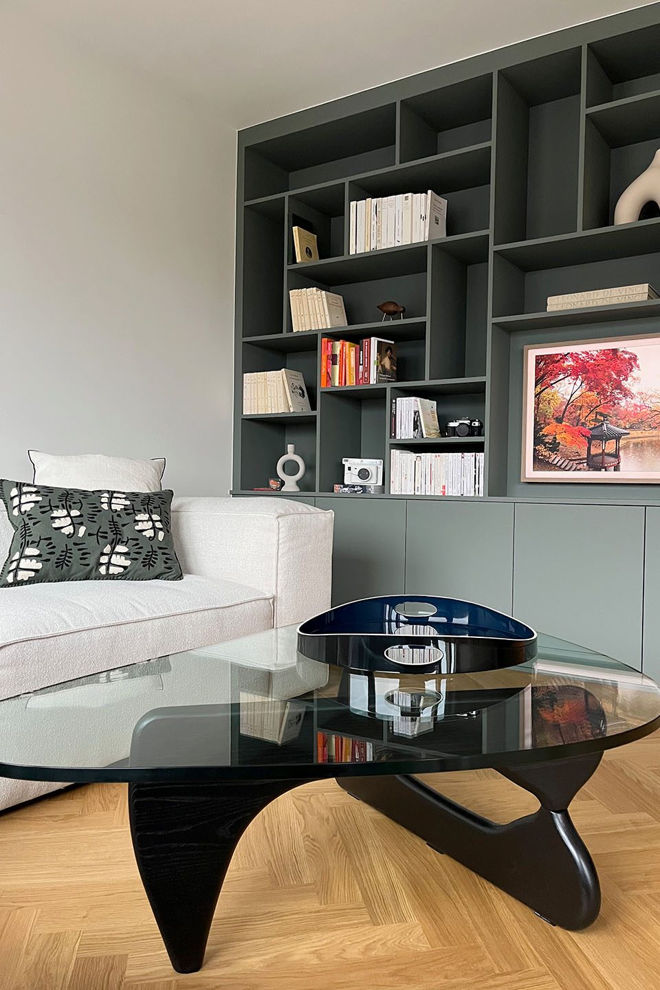 Un salon moderne avec un canapé blanc, une table basse en verre noir, et une étagère grise contenant des livres et des objets décoratifs.