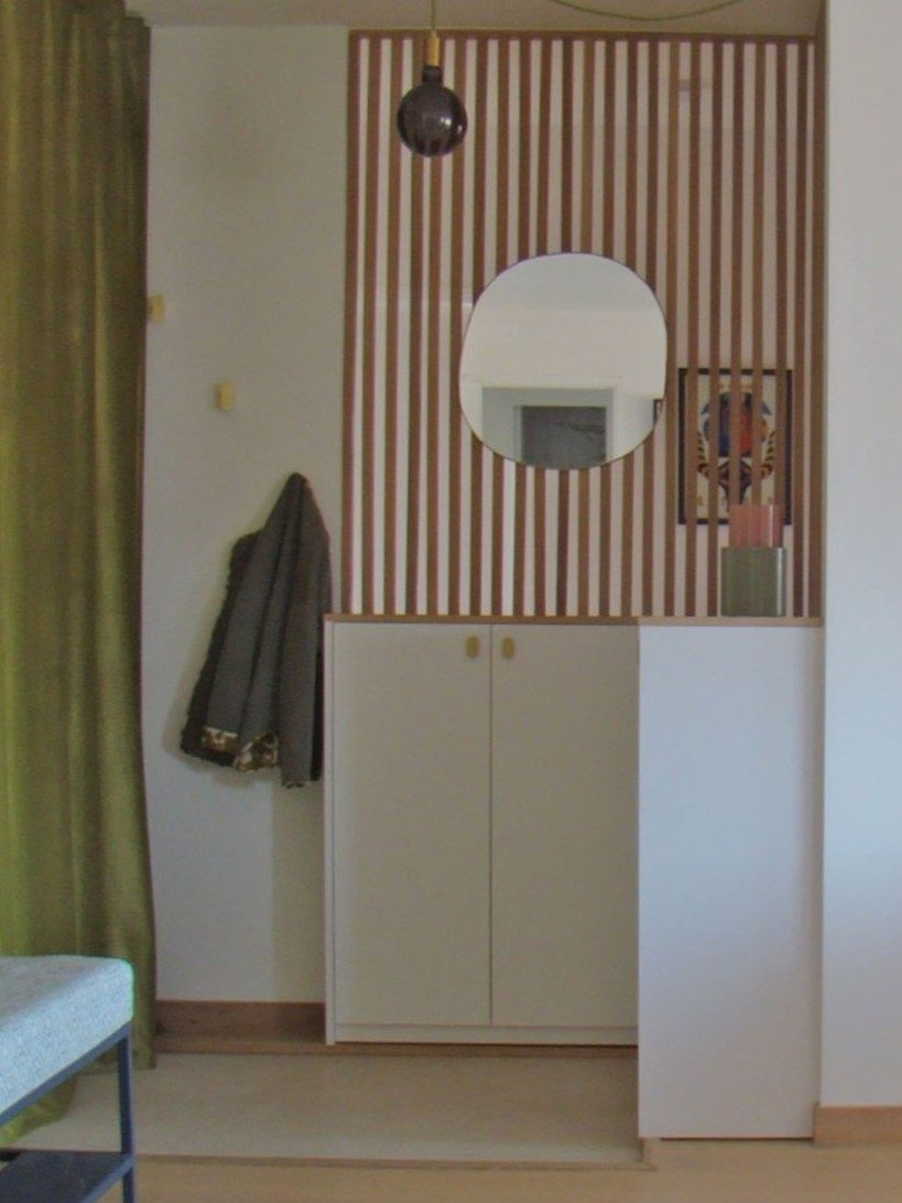 Un coin d'intérieur moderne avec un mur à rayures, un miroir ovale, une penderie blanche, un rideau vert et une lampe suspendue.