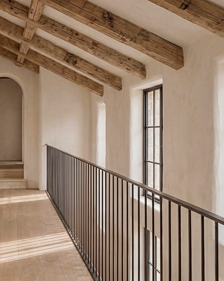 Intérieur d'un couloir avec un plafond en poutres en bois, un garde-corps en métal noir et des fenêtres donnant sur l'extérieur.