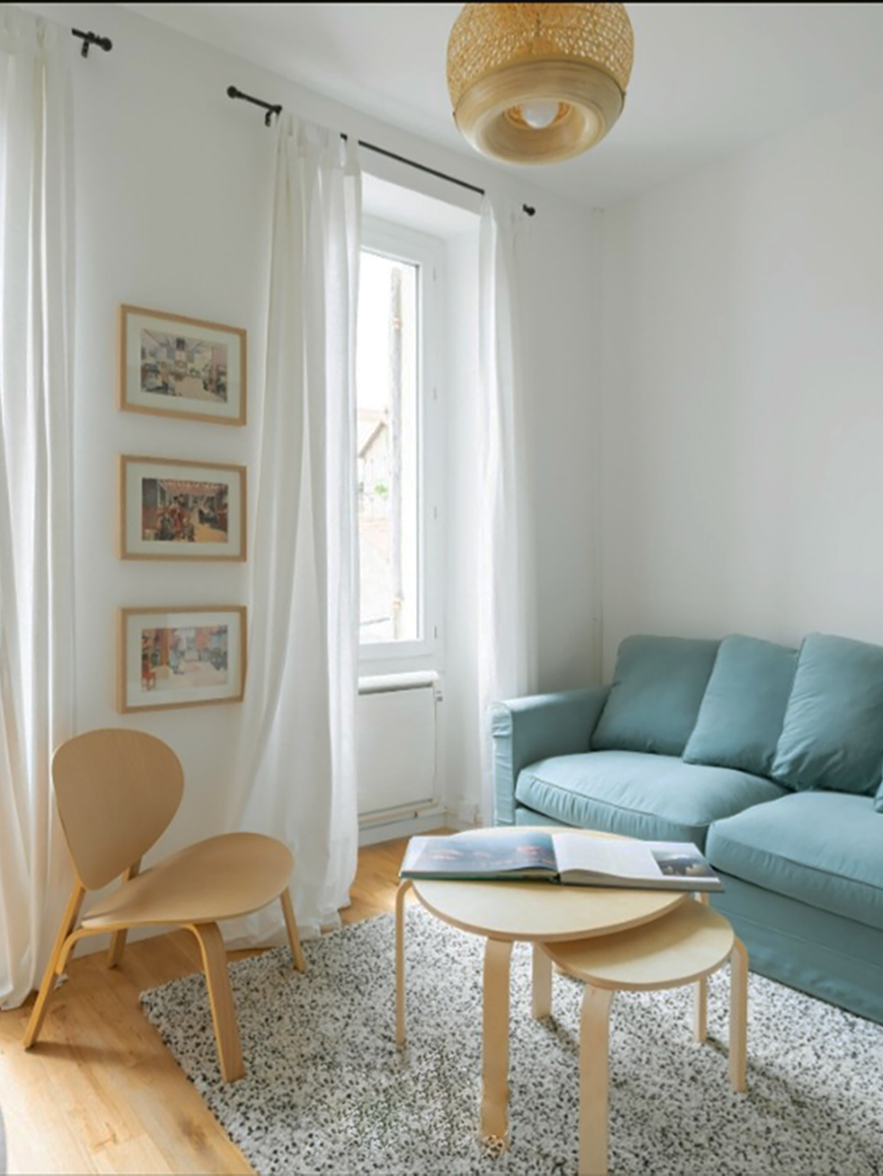 Intérieur d'un salon lumineux avec un canapé bleu, une table basse en bois, une chaise en bois et des murs blancs. Des rideaux blancs ornent la fenêtre, avec des œuvres encadrées accrochées au mur.